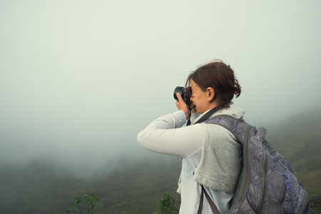 Caucasian woman making photo standing on the top of mountain. It is very cloudy but she tries to shoot.の写真素材