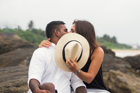 Young mixed race couple kissing sitting on stone. They close faces with hat. Concept of private momentsの写真素材