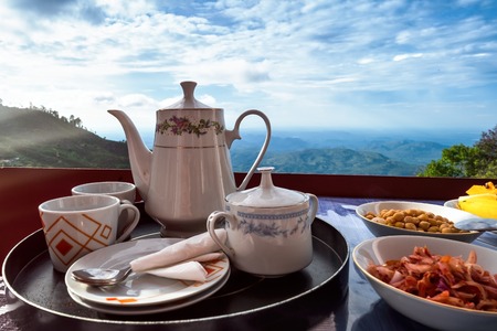 Teapot cups and vegetarian food on table. Asian breakfast with view on mountain and plains. Sunrise in Sri Lankaの写真素材