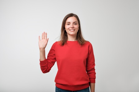 Friendly-looking european teenager dressed in red pulover saying hello, waving her hand. Positive human emotions, feelings or facial expressionsの写真素材