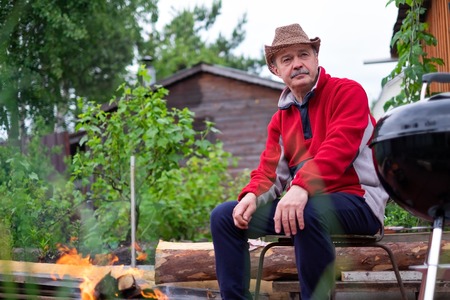 Barbecue time. European man in hat waiting for grilled food. He is sitting near fire and rest.の写真素材