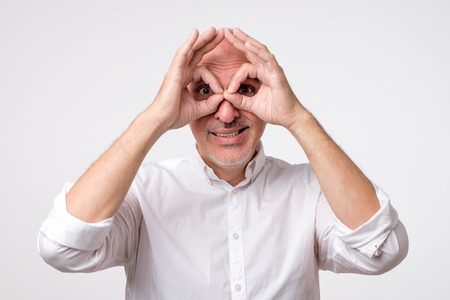 Mature caucasian man showing owl sign with fingers. Making funny grimace. Have a good mood