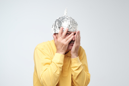 Caucasian mature man in a tin foil hat displeased closing his face with hands. Afraid of radiation or aliensの写真素材