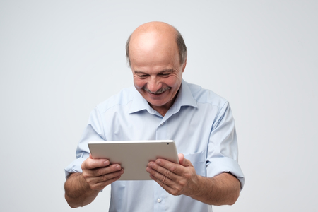 Working on tablet computer. Cheerful mature european man working on digital tablet smiling reading good news.の写真素材