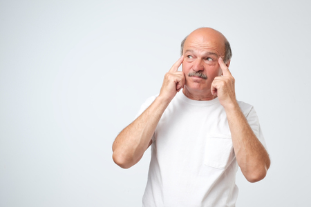 Let me think a little and find sollution. Isolated portrait of puzzled senior male looking up and holding fingers on his temples, trying to remember something or wordの写真素材