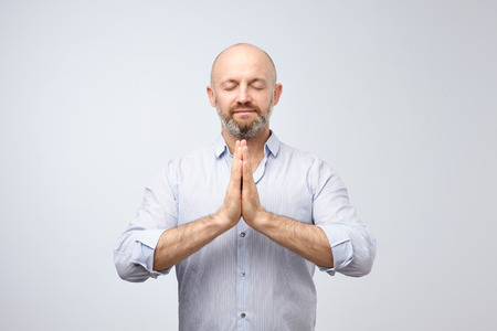 Yoga and meditation at office during work day. Handsome bald man with bristle keeping eyes closed while meditating, feeling relaxed, calm, peaceful holding hands in mudra sign.の写真素材