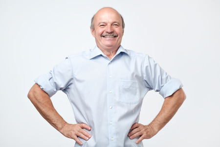 senior handsome man with a proud, satisfied and happy look, with both hands on hips, as if confidently facing a challenge. Akimbo arms pose with a broad smile.の写真素材
