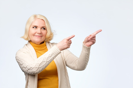 Happy blonde senior woman smiling pointing fingers away, showing something interesting and exciting on studio wallの写真素材