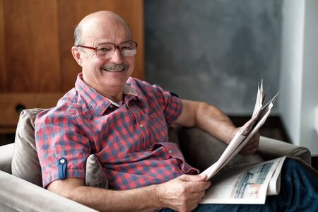 Handsome man sitting in living room, reading newspaper, smiling.