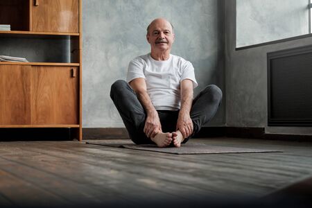 Mature hispanic man sitting barefooted on mat, doing baddha konasana pose sitting in the living room...