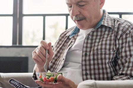 Senior hispanic man sitting in the living room eating fresh vegetable saladの写真素材