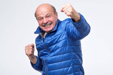 Lucky successful senior man in blue jacket exclaiming, clenching fists, having winning look after...