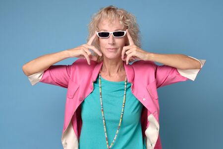 Senior woman wearing stylish sunglasses and smiling at camera on blue wall.の写真素材