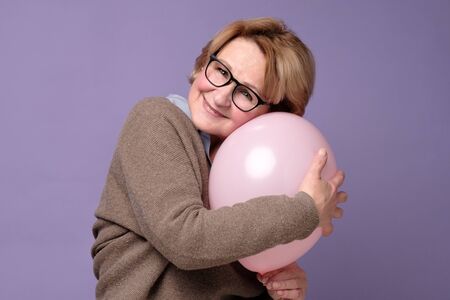 Caucasian senior woman in glasses and sweater hugging pink balloonの写真素材