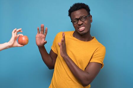 Black young man rejecting to eat apple isolated on blue background