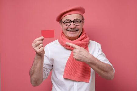 Happy senior hispanic bald man advertising bonus card pointing on it with finger smiling at camera. Guy going to pay by credit card, doing shopping on pink wall.の写真素材