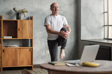 Senior handsome sportman doing stretching and balancing yoga exercise at home using online lesson on notebook. How to stay healthy on quarantine concept.の写真素材