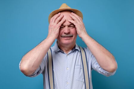 Worried senior man in summer hat suffering from headache. Studio shot on blue wall.