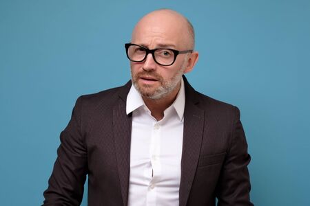 Mature caucasian puzzled male ceo in suit looking at camera over bluebackground
