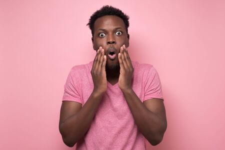 Shocked young african american man in pink clothes looking at the camera in surprise, stunned with interesting story. Human expressions and emotions on studio colored wall.の写真素材
