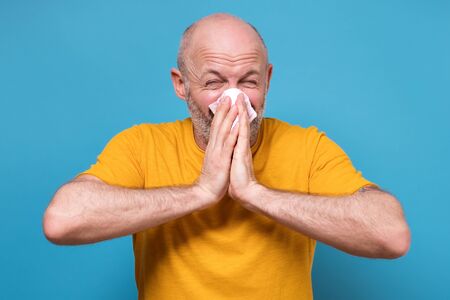 Mature man in yellow clothes sneezing being ill over blue backgroundの写真素材