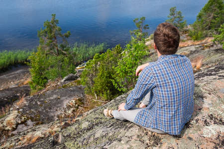 Caucasian man stopping for rest on countryside walk sitting alone on stone near lake.の写真素材