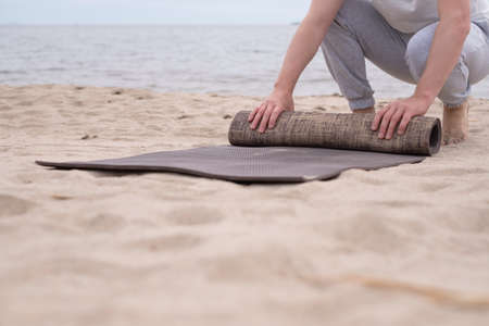 Unrecognizable man rolling up yoga mat after training on sandy beach.の写真素材