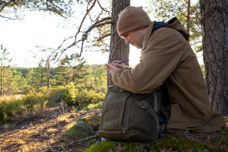 Caucasian man in forest sits on the ground, writing something in diary.の写真素材
