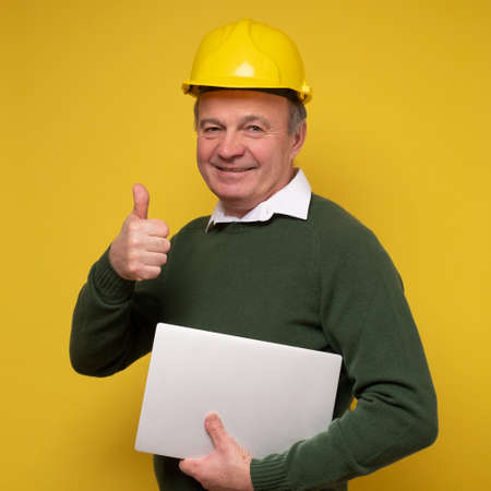 Senior man with special hard hat working using laptop approving showing thumb up