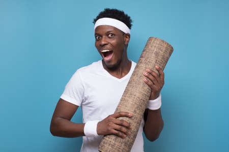 Happy black man holding yoga mat, smiling at camera over blue wall