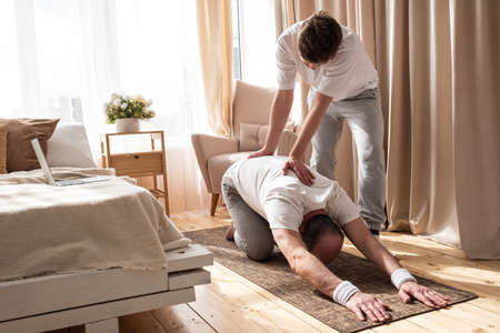Yoga trainer helps to senior man to do yoga asana balsana correctly at home.の写真素材