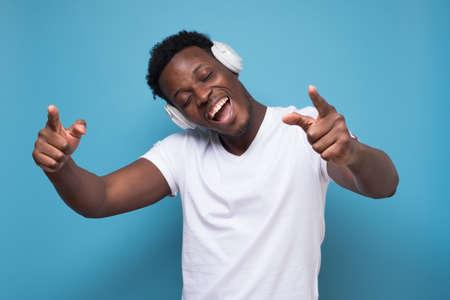 Happy emotive african man listening to music with white earphone. Studio shot on blue wall.の写真素材