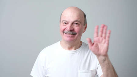 Friendly-looking attractive european pensioner in white t-shirt waives hand in hello gesture while smiling cheerfully. grandfather welcoming his grandson. I am so glad to see you.の写真素材