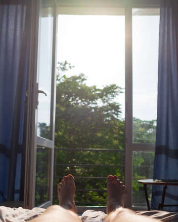 Man legs lying on bed. view on window. It is hot inside without air condition.の写真素材