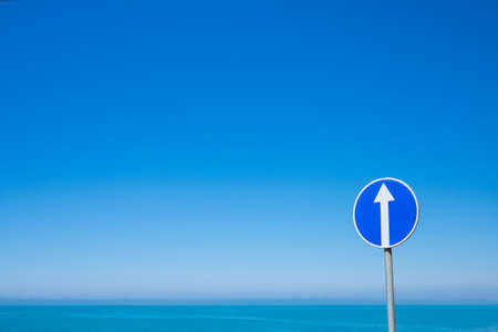 Circle road sign with white arrow on blue sky background. This sign indicates the mandatory direction straight aheadの写真素材