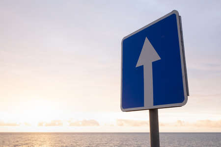 Square road sign with white arrow on blue sky background. It indicates a one way street roadの写真素材