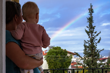 Mother looks at the sky with a baby in her arms, admires the rainbowの写真素材