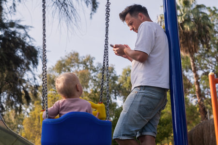 Father with child on a playground looking on phone ignoring his baby swinging.の写真素材