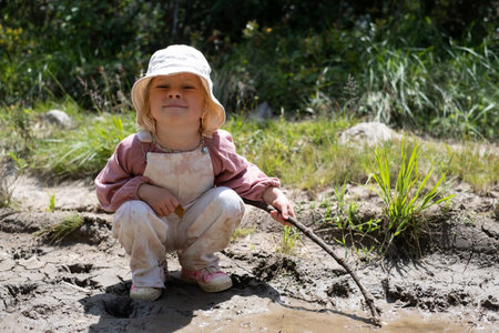 A 3-year-old blonde girl playing in a muddy puddle.