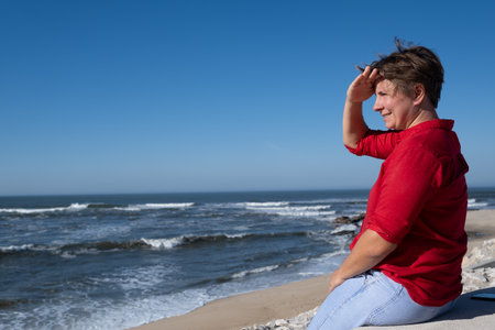 Middle-aged woman sitting on a wall looking at the ocean in Portugal, enjoying a sunny summer day