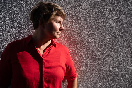 Woman in red shirt standing by the wall, looking aside and smilingの写真素材
