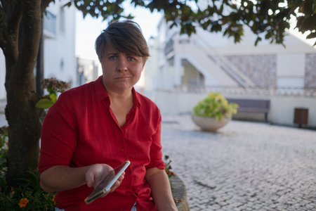 35-year-old European woman sitting outdoors with a phone, not knowing what to doの写真素材