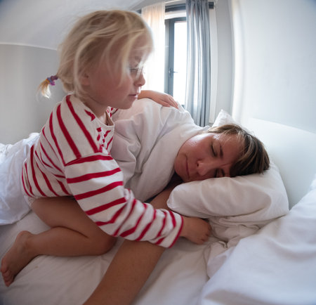 Little girl sitting on the bed trying to wake up her tired sleeping motherの写真素材