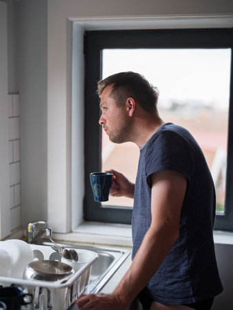 Young man standing thoughtfully by the kitchen window drinking coffee, morning moment of reflectionの写真素材