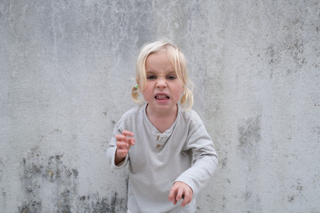 Little 4 Year Old Blonde Girl Standing by a Wall, Pretending to Be Angry and Showing Clawsの写真素材