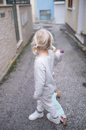Little 4 Year Old Girl Walking Through Narrow Streets with a Scooter Without a Helmetの写真素材