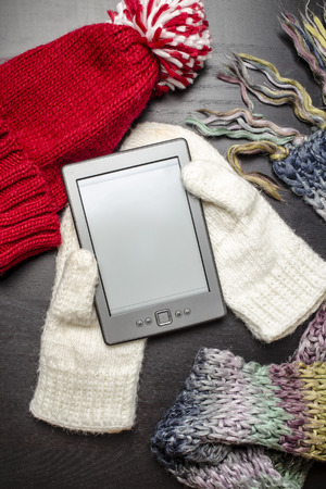 E-book lying on the white gloves, as if lying in the hands of the person near to a black wooden table are other colorful winter clothes: a red hat and scarfの写真素材