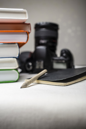 modern digital SLR camera on the table next to a stack of books, and in the foreground a notebook and pencil on a dark backgroundの写真素材