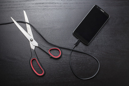 Scissors cut the cable connected to a mobile phone on a dark backgroundの写真素材