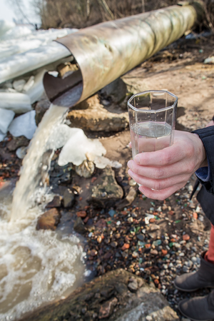 Man with a glass of water in his hand stand near a sewer from which water flows into the river in the spring sunny dayの写真素材